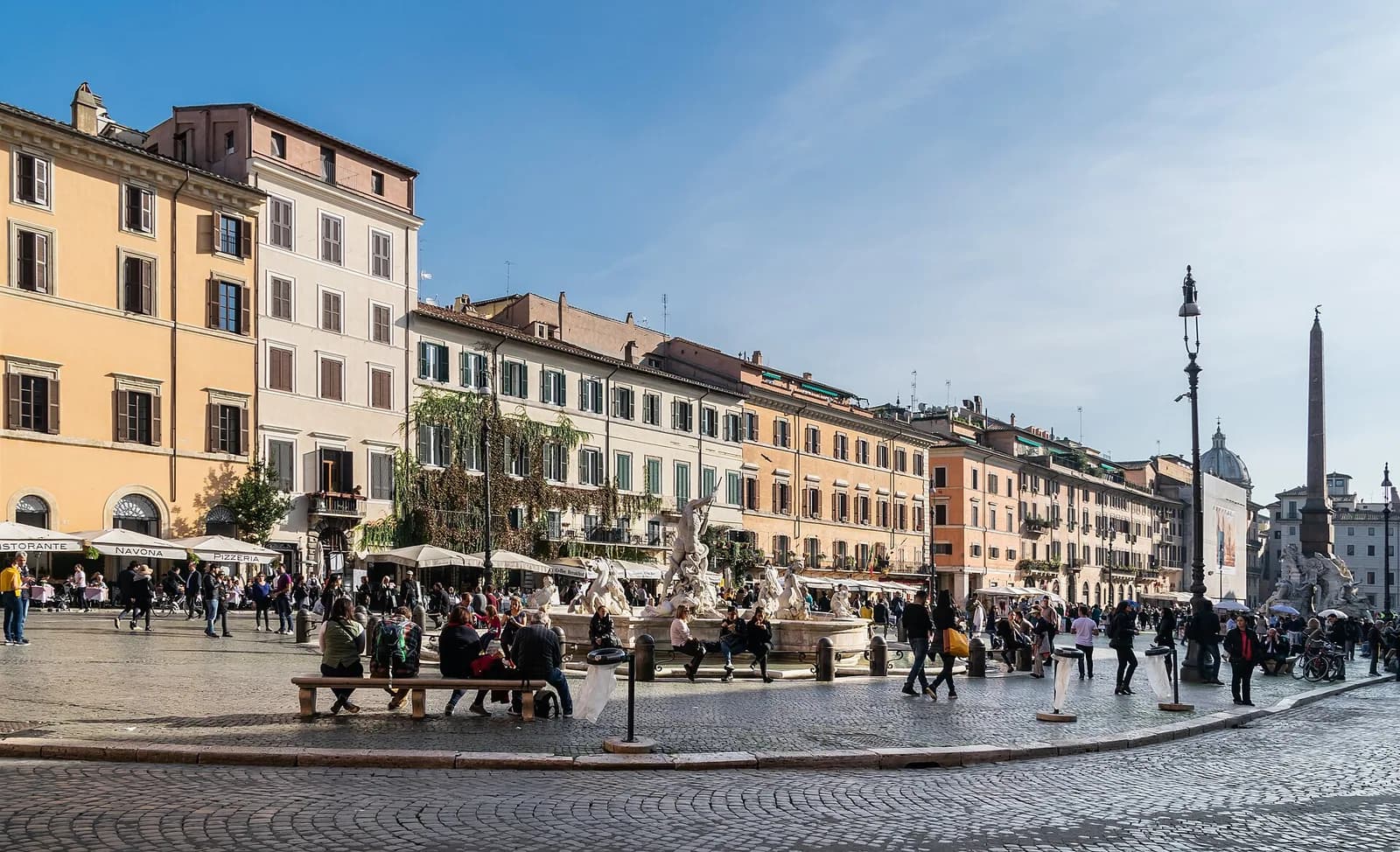 Wide view across Piazza Navona in Rome, with Bernini's Fountain of the Four Rivers and the ochre palazzi lining the square under a clear sky